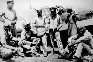 A black-and-white photograph of several African American military pilots gathered casually beside a fighter aircraft marked with a star insignia. The men wear flight suits, caps, and goggles, and appear relaxed as they stand and sit together on the ground near the plane, talking and preparing their gear. The aircraft’s fuselage and cockpit are visible in the background.